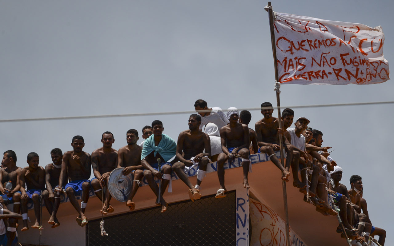 Inmates stand on the prison roof waving a flag that reads "We want peace but don't run away from war" during a rebellion in Alcacuz Penitentiary Center near Natal, Rio Grande do Norte state, northeastern Brazil on January 16, 2017. The latest in a string of brutal prison massacres involving suspected gang members in Brazil has killed 26 inmates, most of whom were beheaded. The bloodbath erupted Saturday night in the overcrowded Alcacuz prison in the northeastern state of Rio Grande do Norte. Similar violence at other jails in Brazil left around 100 inmates dead in early January. / AFP PHOTO / ANDRESSA ANHOLETE