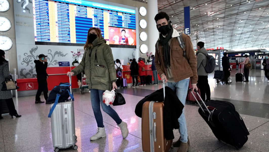 Foreign travellers wearing masks walk past a departures information board at Beijing International Airport in Beijing