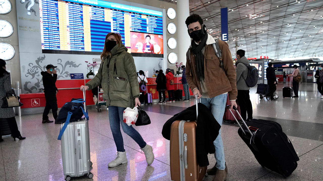 Foreign travellers wearing masks walk past a departures information board at Beijing International Airport in Beijing