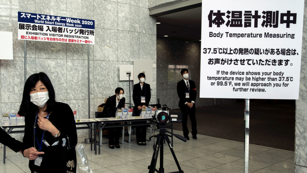 Workers wearing protective face masks, following an outbreak of the coronavirus, stand near a machine measuring body temperature at a convention hall in Tokyo