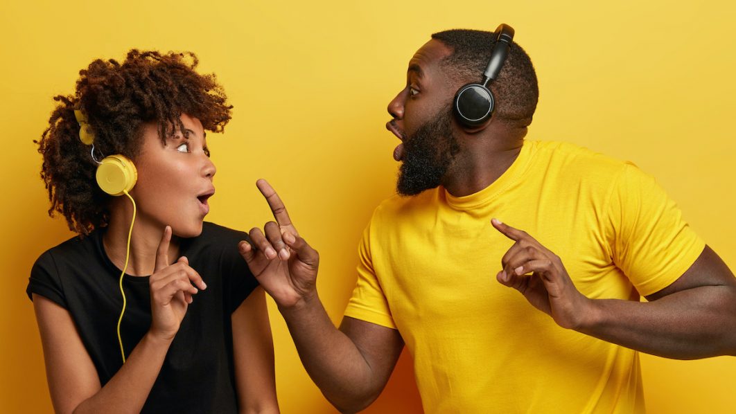 Happy dark skinned students look surprisingly at each other, listen music of same genre, sing favourite melody, listen new composition, filled with positive emotions, wears t shirt, isolated on yellow