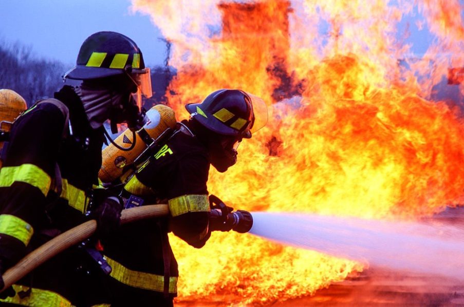 Two firemen spraying water at base of orange flames.
