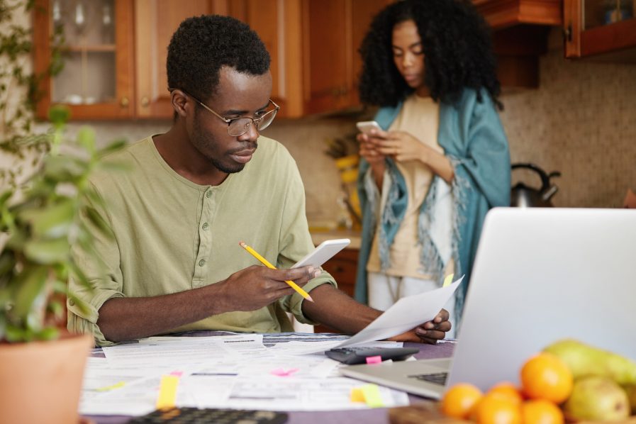 Portrait of confident dark-skinned male holding mobile phone in one hand and piece of paper in other, dialing number of utility services, trying to solve problem of power cut in his apartment