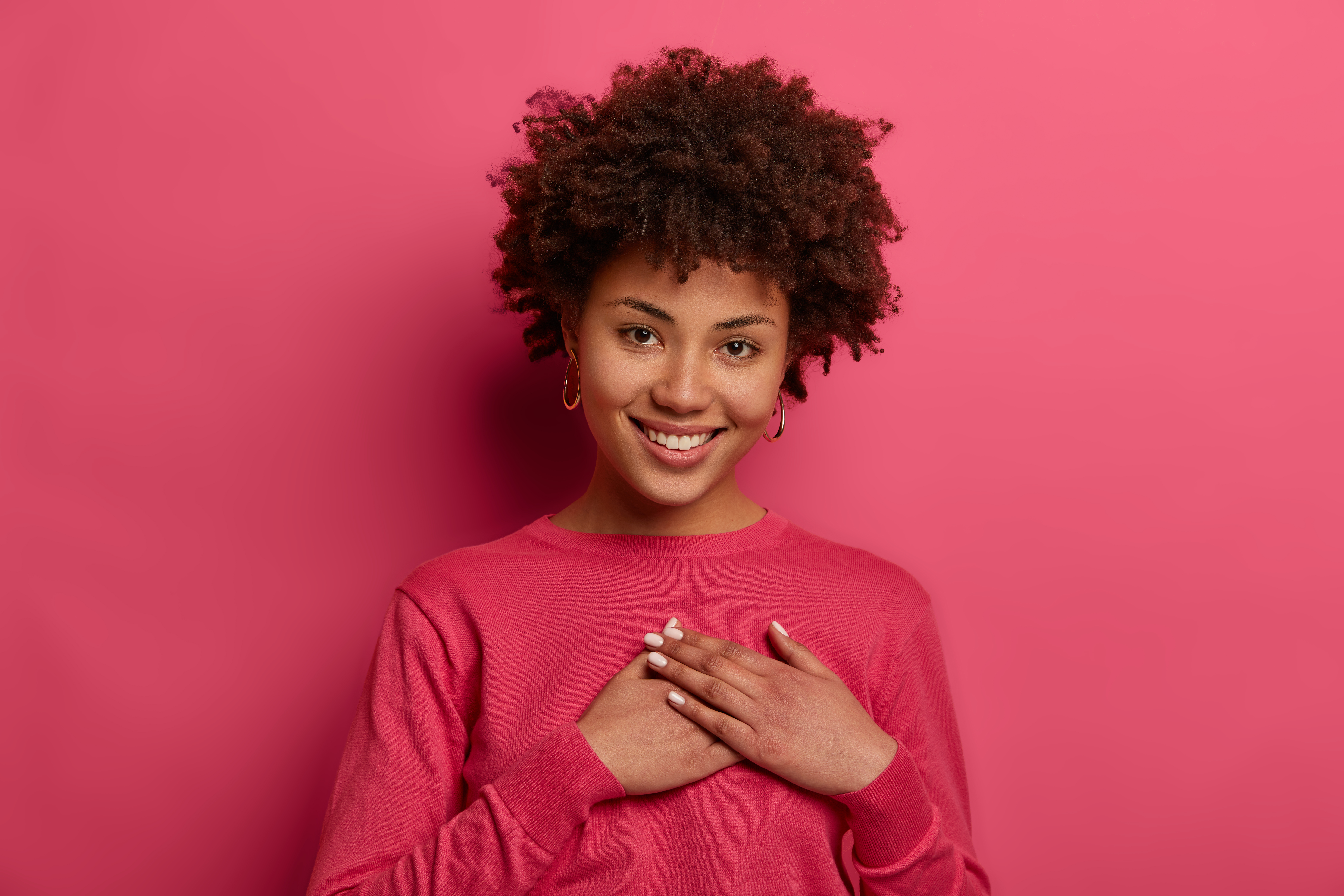 Portrait of touched lovely woman touches heart, makes sign of affection and gratitude, has gentle smile, wears crimson jumper, appreciates nice words, looks gratefully, isolated on pink wall