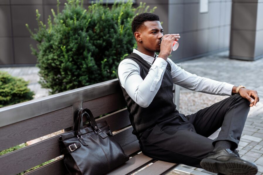 A man seating and taking a drink with bag by the side. Photo from Thirdman in Pexels