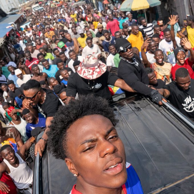 Large crowd surrounds a young male content creator standing through a vehicle sunroof during a street appearance in Liberia.