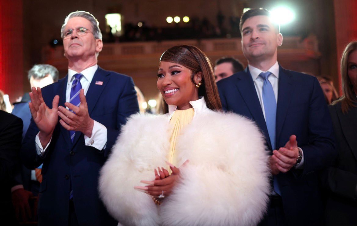 Nicki Minaj smiles at a public political event wearing a white fur coat, standing between two men as an audience applauds behind her.