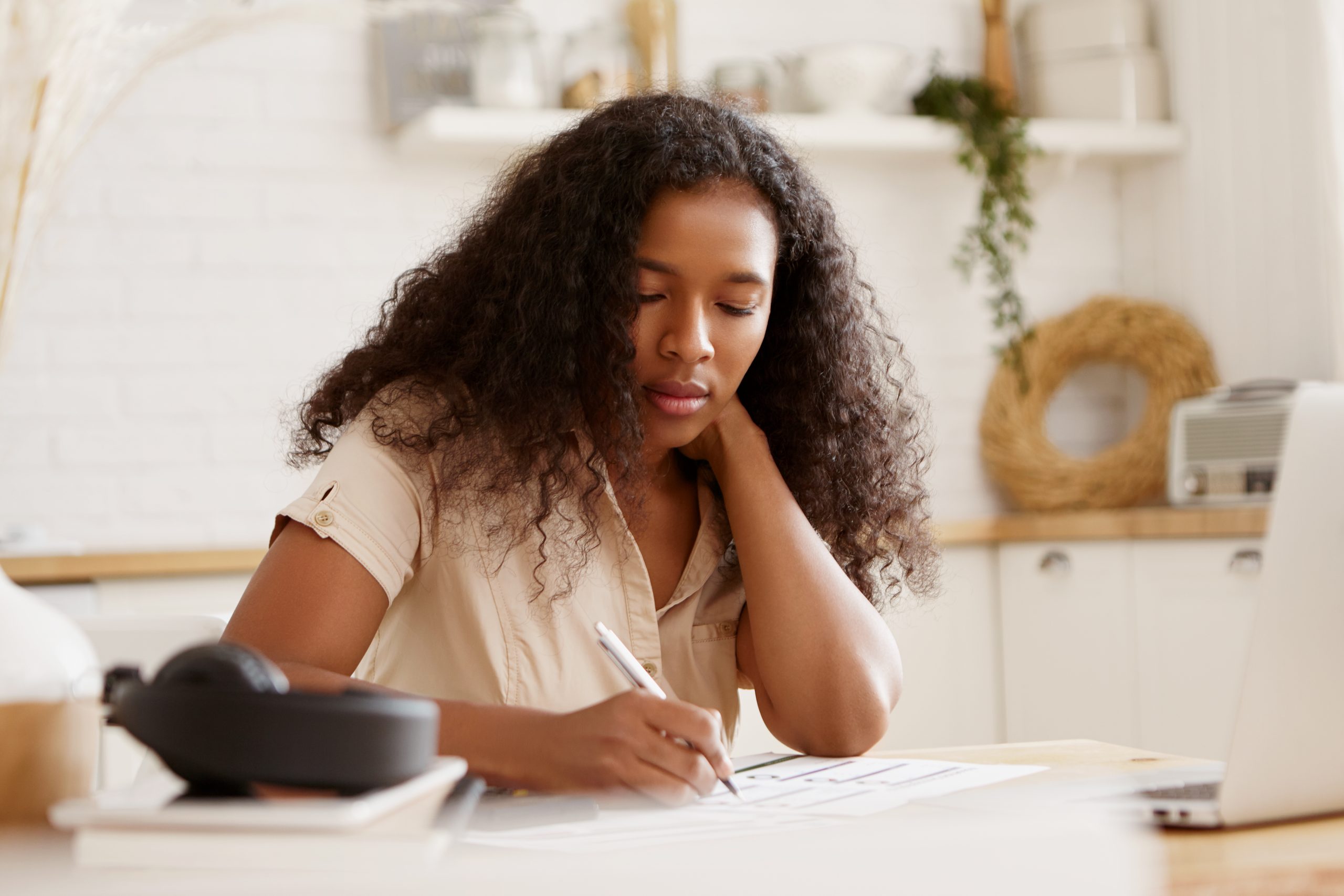 Portrait of concentrated serious African American student girl holding pencil, writing down, preparing for exams or doing homework in kitchen, sitting at dining table with open laptop and books. Photo generated using Canva