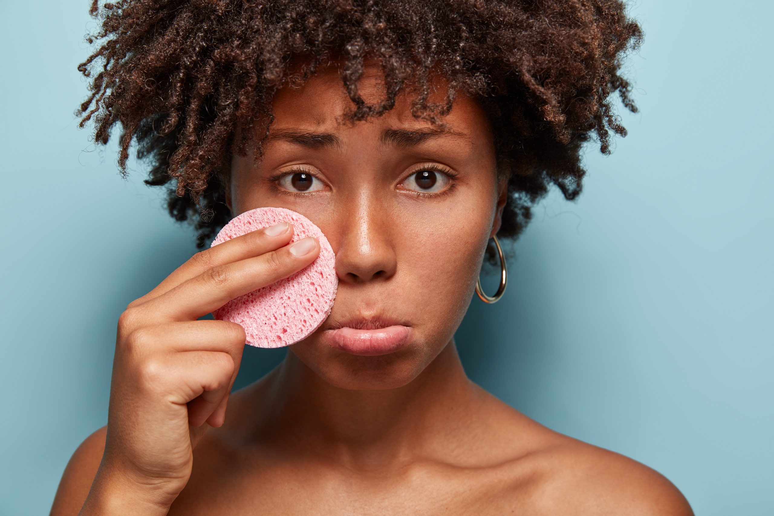 Close up shot of displeased dark skinned woman holds sponge on cheek, purses lips, applies cream on face, poses half nude at camera against blue background. Facial treatment and beauty concept
