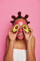 Beauty treatment concept. Happy dark skinned female covers eyes with avocado halves, smiles broadly, applies face mask for rejuvenation and freshness, wrapped in towel, isolated on pink background