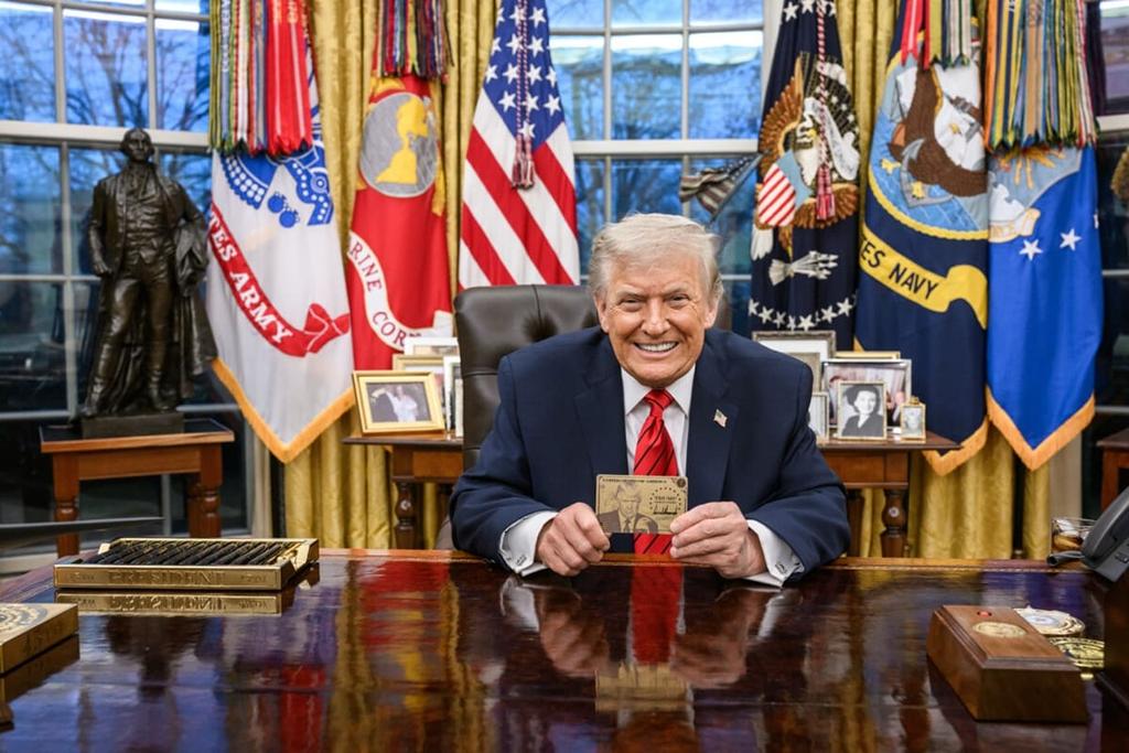 The US president sits behind a desk in the Oval Office holding up a gold-coloured card, with national flags displayed behind him