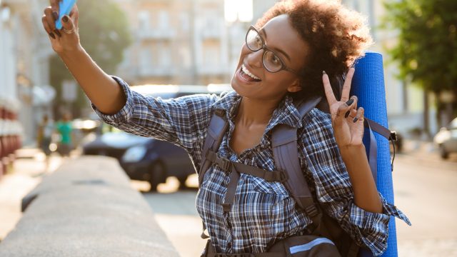 Young beautiful african female traveler with backpack smiling, making selfie.