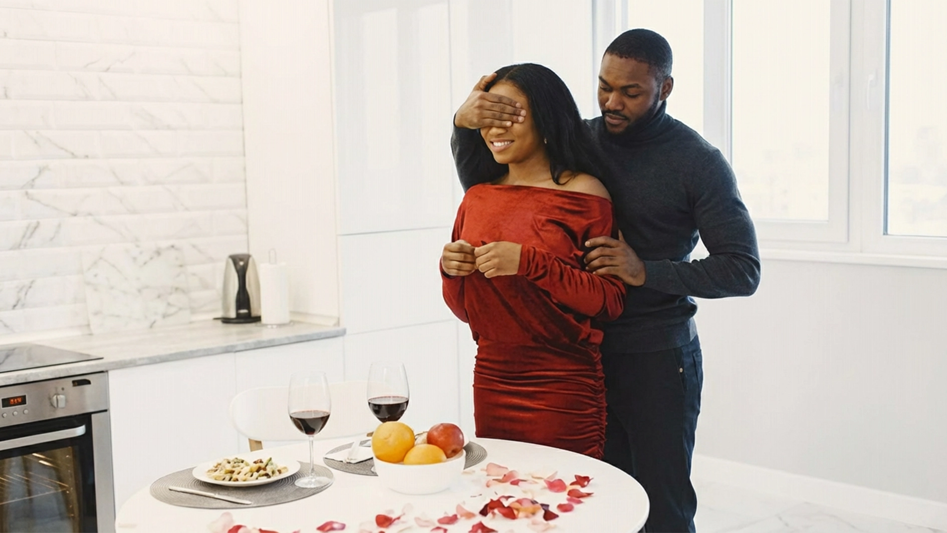 Valentine’s Day surprise dinner at home with a couple, wine glasses, food, and rose petals on the table.