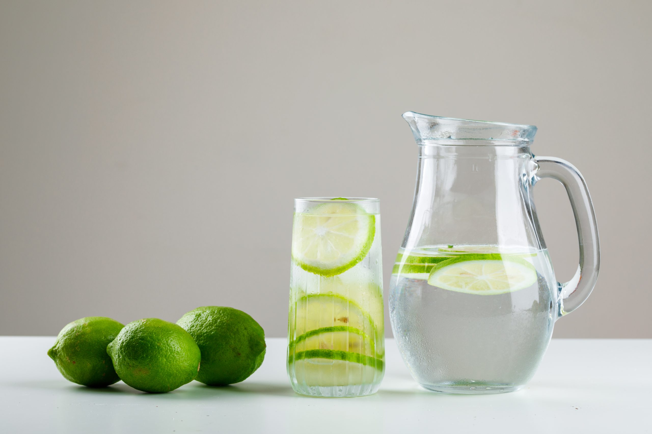 Lemonade with lemons in glass and jug on white and grey background, side view.
